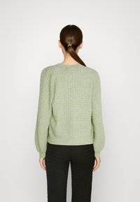 Grön texturerad tröja med rundad halsringning, långa ärmar och ribbat nederkant, ihop med svarta texturerade byxor.