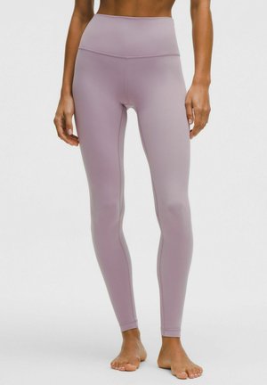 Kobieta w wysokim stanem, bezszwowych legginsach w kolorze mauve, stojąca boso na jasnym, jednolitym tle.