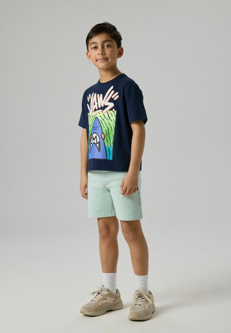 Garçon debout en short bleu clair, T-shirt bleu marine avec un motif requin coloré, chaussettes blanches et baskets beiges sur fond uni.