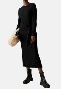 Robe en tricot côtelé noire avec des manches longues et un détail de nouage sur le côté. Associée à des bottes de cheville à fermeture éclair robustes noires. Sac texturé beige à la main.