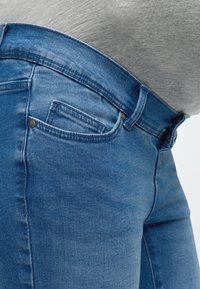 Gros plan sur une personne portant un jean en denim bleu moulant et un t-shirt gris, montrant la ceinture et les détails de la poche avant.