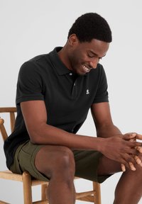 Black polo shirt with short sleeves, featuring a small logo on the chest, paired with green shorts, sitting on a wooden chair.