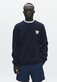 Pull bleu marine avec poignets et ourlet côtelés, arborant un logo "W" crème sur le devant, porté avec un short bleu marine. Texture tricotée lisse.