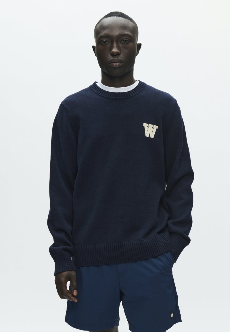 Pull bleu marine avec poignets et ourlet côtelés, arborant un logo "W" crème sur le devant, porté avec un short bleu marine. Texture tricotée lisse.