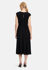 Schwarzes Midi-Kleid mit kurzen Ärmeln, einer Schlüsselverschlussöffnung im Rücken und einem ausgestellten Saum, aus strukturiertem Gewebe gefertigt. Kombiniert mit schwarzen hohen Sandalen.