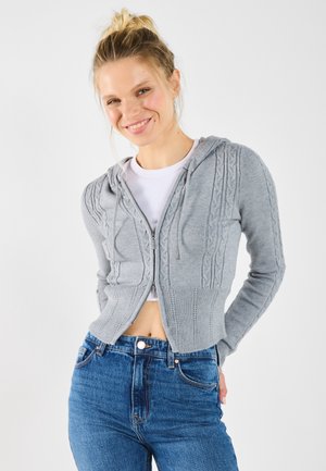 Gray zip-up cardigan with cable knit design, hood, and ribbed hem; worn over a white t-shirt with high-waisted blue jeans.