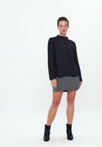 Schwarzes Oberteil mit Polka-Dots, langen Ärmeln und rundem Ausschnitt, kombiniert mit einem grauen Minirock. Das Model trägt schwarze Knöchelstiefel. Einfaches, modernes Design.