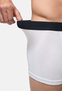 Eine Person zieht den schwarzen Bund weißer Boxershorts von der Taille weg und zeigt dabei die elastische Dehnung und den Markennamen "INDICODE."