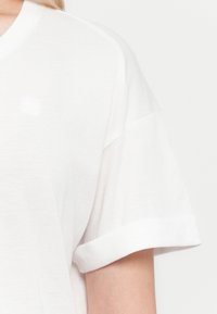Close-up of a person wearing a white short-sleeve t-shirt with a folded cuff and a subtle logo on the chest.