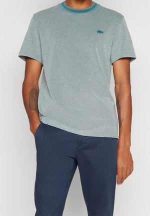 Homme portant une chemise bleu clair à manches courtes avec un logo brodé et un pantalon bleu marine, une main dans la poche, sur fond uni.
