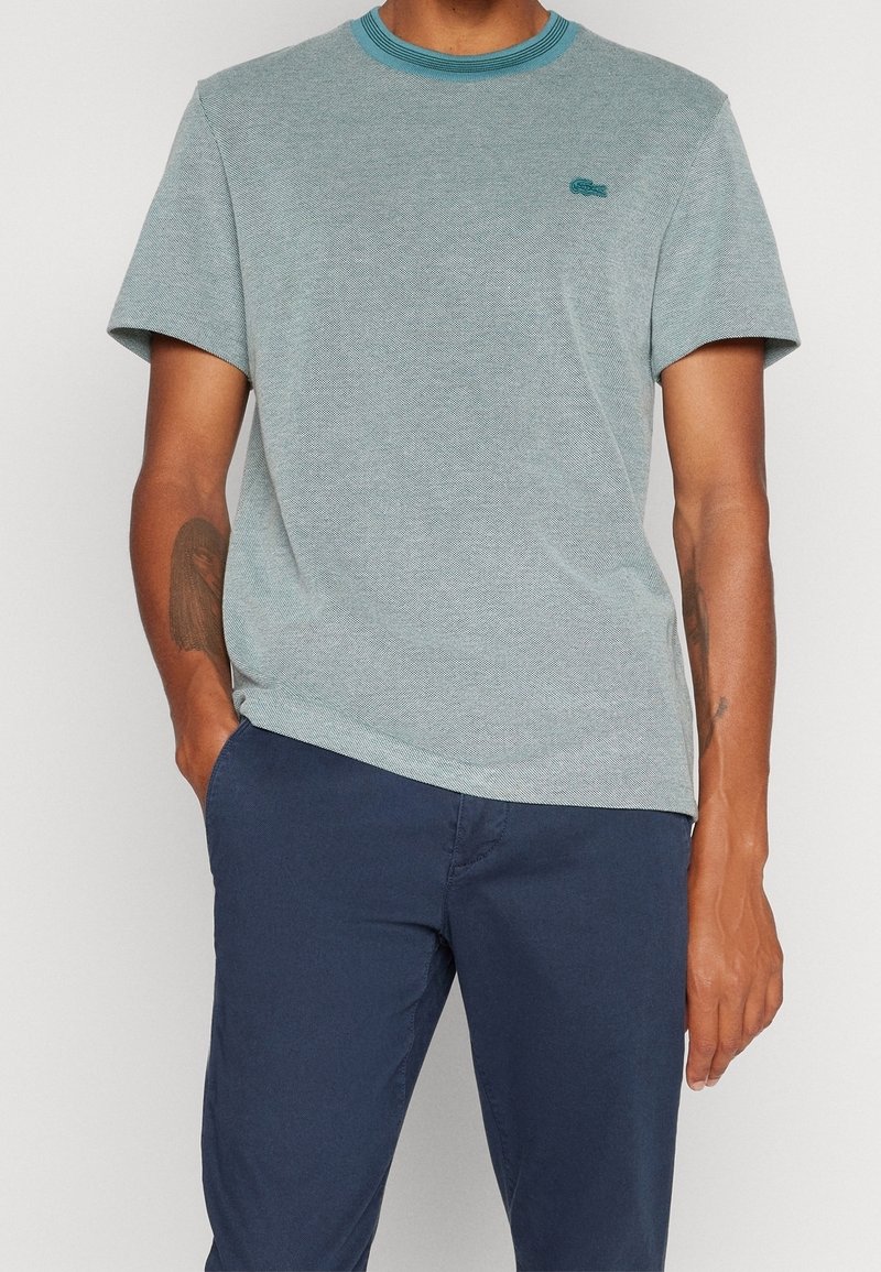 Homme portant une chemise bleu clair à manches courtes avec un logo brodé et un pantalon bleu marine, une main dans la poche, sur fond uni.