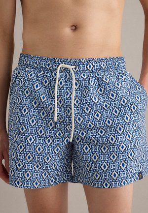 Mann trägt blaue und weiße Schwimmshorts mit geometrischem Muster, weißem Kordelzug und seitlichen Taschen.