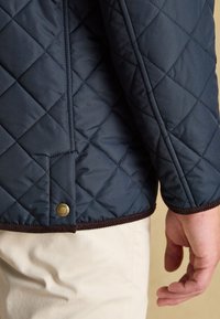 Marineblaue gesteppte Jacke mit Diamantsteppung und braunem Besatz, getragen über beigen Hosen, zeigt den unteren Rücken und die entspannte rechte Hand.