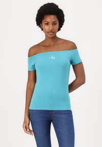 Top turquesa sin hombros hecho de tela elástica, con mangas cortas y un pequeño logo blanco en el pecho, combinado con jeans azules.