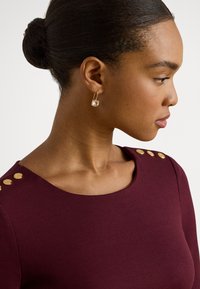 Vīnsarkans tops ar apaļu izgriezumu, uz pleca ar zelta pogu rotājumiem. Modelis valkā zeltu auskaru ar caurspīdīgu dārgakmeni.