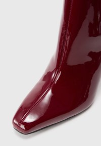 Bota de tornozelo de couro patente bordô com bico fino, design elegante, fecho lateral e acabamento liso e brilhante.