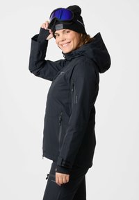 Woman in black winter jacket, beanie, and ski goggles, smiling and adjusting goggles against plain light background.