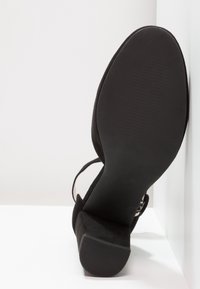Chaussure basse en daim noir pour femme vue de la semelle, avec un bout arrondi, un talon bloc et une bride à la cheville.
