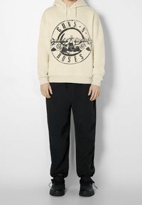Sudadera beige con un diseño gráfico negro que presenta "GUNS N ROSES", combinada con pantalones negros y zapatillas negras. Ajuste casual y relajado.