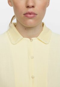 Blusa giallo pallido con colletto arrotondato, chiusura a bottoni e delicati punti ondulati lungo il bordo del colletto; tessuto leggero.