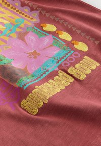 Tissu rose avec motif floral vibrant brodé, bordure décorative bleue, formes de canards jaunes et texte jaune texturé sur fond rouille.
