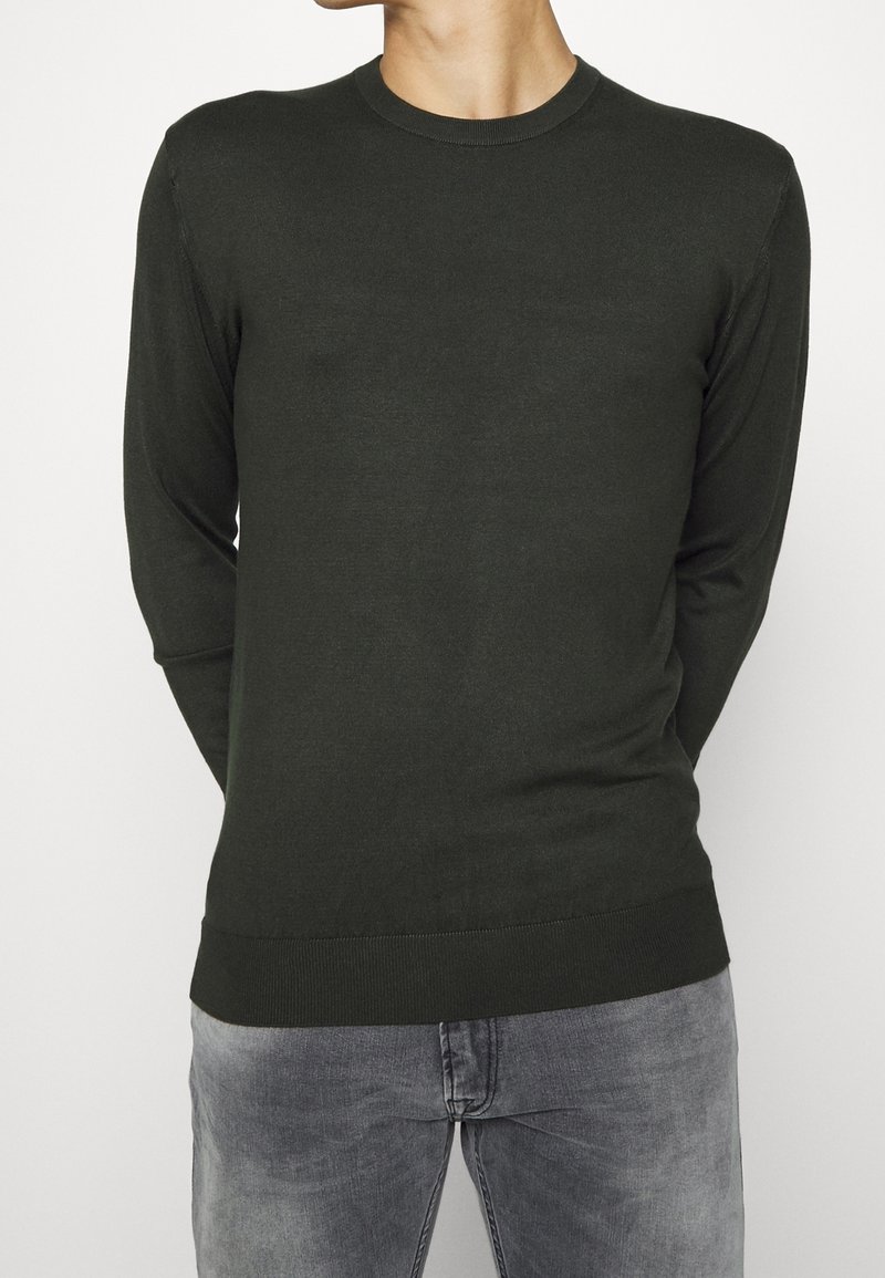 Uomo che indossa un maglione a maniche lunghe verde scuro semplice e jeans grigi sbiaditi, in piedi con le mani dietro la schiena su uno sfondo bianco.