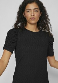 Čierne šatstvo s textúrovaným povrchom, ktorý obsahuje prešívaný vzor a nadýchané krátke rukávy. Okrúhly výstrih, voľný strih a jemná tkanina.