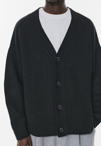Cardigan negru tricotat cu decolteu în V, manșete și tiv ribbate, având cinci nasturi pe față, suprapus peste un articol vestimentar gri.