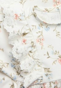 Vit tyg med blommönster i rosa, blått och brunt. Volangspets längs kanten. Enkel halsringning och dämpad, mjuk textur.