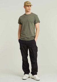 Olijfgroene t-shirt, korte mouwen, ronde hals; zwarte cargobroek met zijzakken; beige pet, witte sneakers met zwarte accenten.