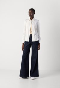 Helles, langärmliges Knopfhemd über einem beigen Oberteil, kombiniert mit weit geschnittenen dunkelblauen Jeans und weißen Sneakers.