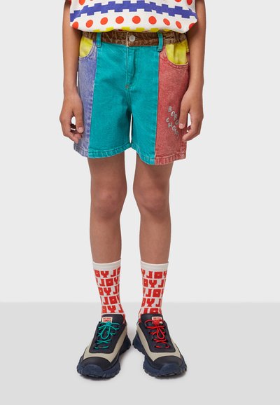 Shorts en denim multicolores avec des panneaux bleus, verts, violets et rouges, dotés de poches jaunes ; associés à des chaussettes à motifs et des baskets chunky.