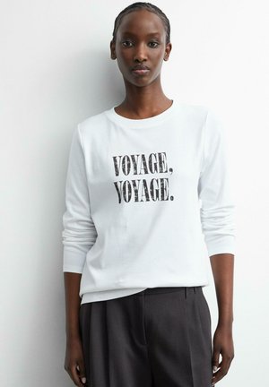Frau trägt ein weißes Langarmshirt mit schwarzem Text "VOYAGE, VOYAGE.", kombiniert mit dunklen Hosen, steht vor einer weißen Wand.