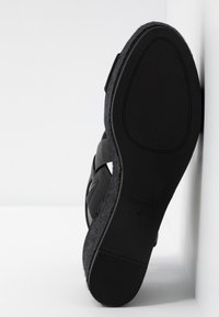 Sandale compensée noire avec semelle texturée et larges brides, taille 38, positionnée debout contre un coin de mur et de sol blanc.