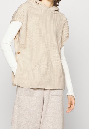 Persona che indossa un poncho beige lavorato a maglia senza maniche con cappuccio sopra una maglia bianca a maniche lunghe e pantaloni beige a gamba larga.
