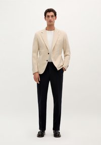 Blazer de lino beige con solapas de muesca y dos botones, combinado con una camiseta de cuello redondo blanca y pantalones oscuros en color navy, acentuado con zapatos negros.