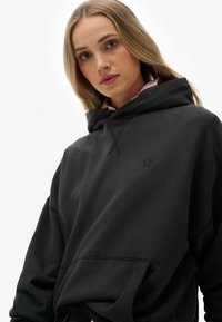 Sudadera con capucha negra fabricada en tejido suave, con un bolsillo frontal, puños acanalados y un discreto detalle de logo en el pecho.