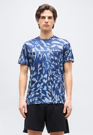 Tânăr în picioare, purtând un tricou Champion albastru cu model și pantaloni scurți negri, pe un fundal simplu, deschis la culoare.