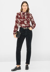 Blouse met lange mouwen in een gedurfde rood, wit en zwart geometrisch patroon, gecombineerd met zwarte corduroy broek en zwarte instappers. Zwarte handtas met gouden accenten.