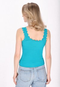 Turquoise ribbel tanktop met golvende randen en een getailleerd ontwerp, gecombineerd met lichtblauwe spijkerbroeken. Focus op textura en kleur.