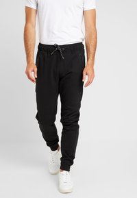 Zwarte joggingsbroek gemaakt van zachte stof met een koord in de taille, voorzien van ritszakken en een contrasterend paneelontwerp. Gemaakt in combinatie met witte sneakers.