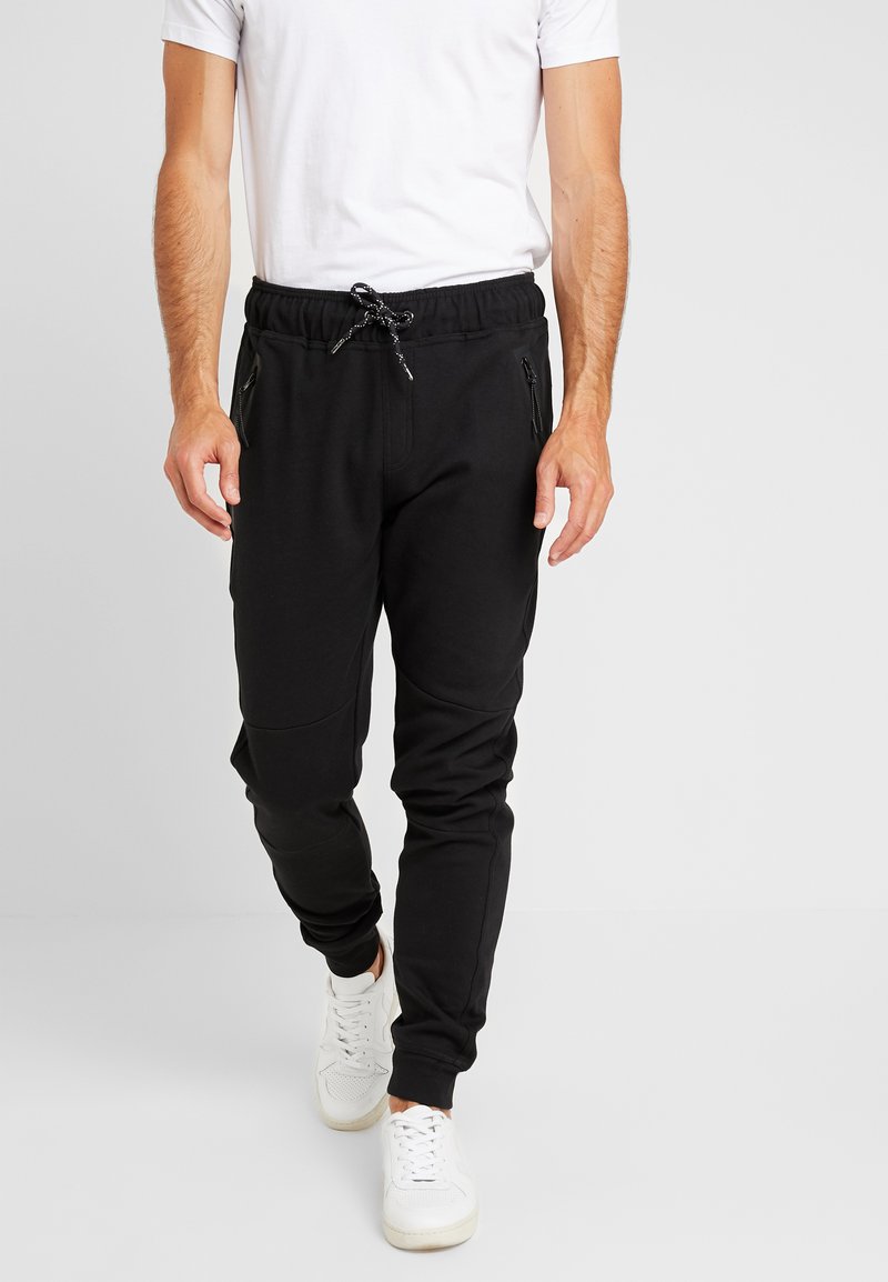 Zwarte joggingsbroek gemaakt van zachte stof met een koord in de taille, voorzien van ritszakken en een contrasterend paneelontwerp. Gemaakt in combinatie met witte sneakers.
