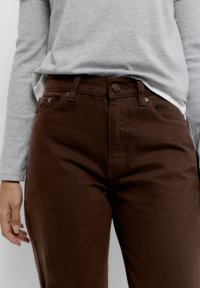 Közeli kép egy sötétbarna nadrágot és világosszürke hosszú ujjú inget viselő személyről, akinek az egyik keze laza tartásban van az oldala mellett.