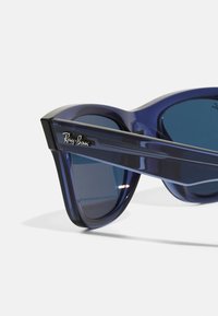 Ochelari de soare Ray-Ban cu o ramă transparentă de culoare albastru închis, lentile întunecate și un logo argintiu pe lateral. Design geometric de tip cat-eye.