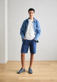 Veste en denim, t-shirt blanc, shorts en denim bleu et baskets grises avec des accents oranges; debout sur un sol en bois contre un mur gris.
