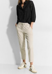 Svart perforerad långärmad skjorta med krage, tillsammans med beige byxor med elastisk midja och vita sneakers med svarta detaljer.