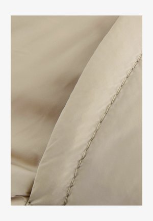 Tessuto beige con una superficie liscia, caratterizzato da un bordo cucito. Il materiale sembra leggero e flessibile, adatto per capi esterni.