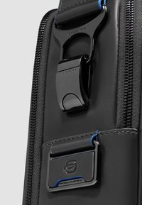 Bolso sintético negro con textura suave, herrajes metálicos, una cremallera prominente y detalles azules en la correa y la etiqueta.