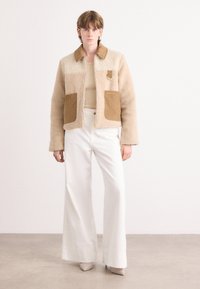Veste en fausse fourrure beige avec des accents en velours côtelé marron et des poches. Assortie à un pantalon large blanc et un haut beige ajusté.