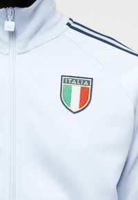 Hvid sportjakke med lynlås og sorte striber på skulderen samt et Italia-emblem med det italienske flag på brystet.